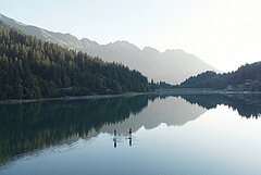 Two people stand-up paddling on a calm mountain lake with forested slopes and mountains reflected in the clear water.