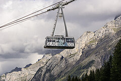  Aerial cable car with a Glacier3000 sign suspended before a backdrop of mountains in summer and a cloudy sky.