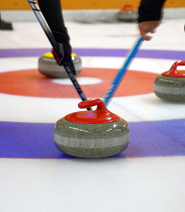 Two curling players swipe a red and grey curling stone into a blue, white and red ring, with three more curling stones next to it.
