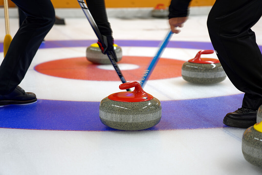 <p>Zwei Curlingspieler wischen einen rot-grauen Curlingstein in den blau-weiss-roten Ring. Daneben liegen drei weitere Curlingsteine.</p>