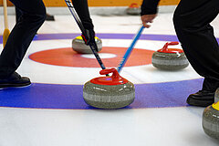 Zwei Curlingspieler wischen einen rot-grauen Curlingstein in den blau-weiss-roten Ring. Daneben liegen drei weitere Curlingsteine.
