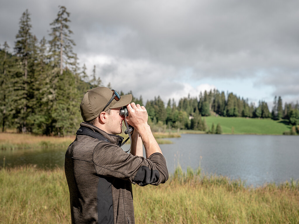 <p>Ein Ranger, welcher durch ein Fernglas schaut. Im Hintergrund ist der Lauenensee und Bäume zu sehen.</p>