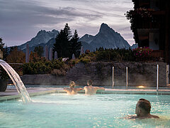 Three people swim in an outdoor whirlpool with mountains in the background.