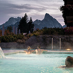 Drei Personen schwimmen in einem Whirlpool im Freien, mit Bergen im Hintergrund.