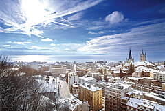 Verschneite Dächer einer Stadt mit Kathedrale und Stadtzentrum, im Hintergrund der Genfersee unter winterlichem Himmel.
