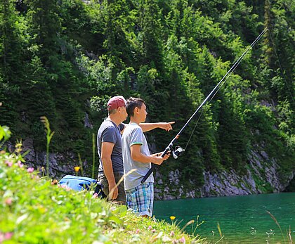 2 Personen stehen am Ufer eines Sees auf der Wiese und fischen