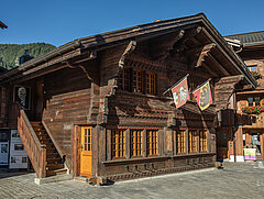 You can see a small wooden chalet with two floors. On the first floor, two flags hang by the windows. One is the Bernese flag with two corners in red and a yellow stripe in the centre where the black bear stands. The second is the Saanen flag - red background and a white crane standing on three grey stones. The chalet has small windows and an outside staircase leads to the first floor.