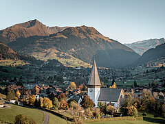Das Dorf Saanen an einem frühen Herbstmorgen. Die Kirche ist bereits im Sonnenlicht, während das Dorf noch im Schatten liegt. Im Hintergrund sieht man Giferspitz und Wasserngrat in herbstlichen Brauntönen.