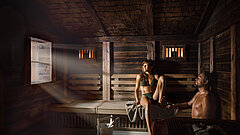 Une femme et un homme sont assis dans un sauna. Au premier plan, un poêle et sur le sol, une chaudière à bois.
