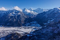 Luftaufnahme einer verschneiten Alpenlandschaft mit Talboden, steilen Berghängen und markanten Gipfeln im Winterlicht.