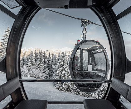 Das Foto stammt aus einer modernen 10er-Gondelbahn. Die Gondelbahn hat schwarze Lederbänke und viel Glas. Das Design ist oval und wirkt futuristisch. Ausserhalb der Gondel kreuzt eine andere Gondel mit zwei Personen darin. Draussen ist die Landschaft verschneit und nur auf den Bergspitzen sieht man die Sonne.