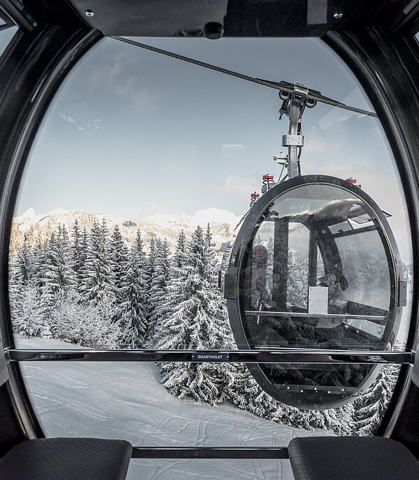 Das Foto stammt aus einer modernen 10er-Gondelbahn. Die Gondelbahn hat schwarze Lederbänke und viel Glas. Das Design ist oval und wirkt futuristisch. Ausserhalb der Gondel kreuzt eine andere Gondel mit zwei Personen darin. Draussen ist die Landschaft verschneit und nur auf den Bergspitzen sieht man die Sonne.