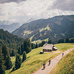 Three cyclists ride down the narrow road from the Mittelbergpass into the Grischbachtal. In the background, a mountain panorama stretches beneath dramatic clouds.