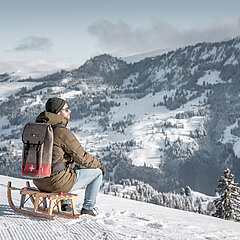 Person sitzend auf einem Holzschlitten vor verschneiter Bergkulisse.