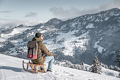 Person sitting on a wooden sled in front of a snowy mountain backdrop.