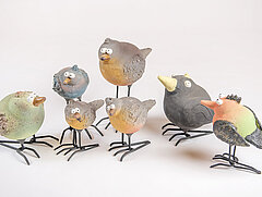 Sieben bunt bemalte Vögel aus Ton mit schwarzen Füssen aus Metall auf weissem Hintergrund.