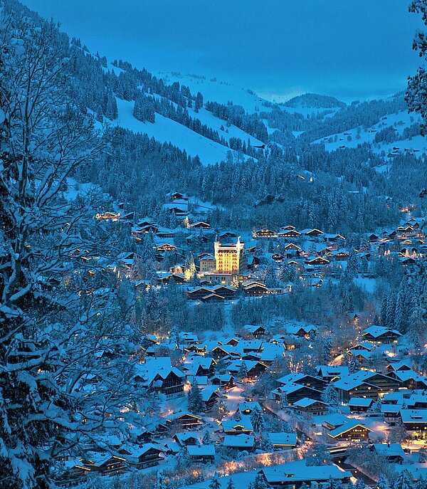 View of the village of Gstaad in the snow, with the illuminated Gstaad Palace and mountains in the background.