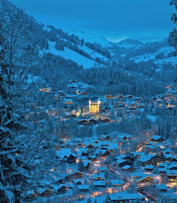 <p>Blick auf das verschneite Dorf Gstaad mit dem beleuchteten Gstaad Palace und Berge im Hintergrund.</p>