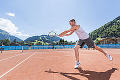 Ein Mann in sportlicher Bekleidung steht auf einem Tennisplatz und spielt den Tennisball zu seinem Mitspieler.