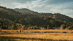 Landschaft mit grüner Wiese im Vordergrund, einem ruhigen See in der Mitte und bewaldeten Hügeln im Hintergrund unter blauem Himmel mit vereinzelten Wolken. Zwei Personen stehen am Ufer des Sees.