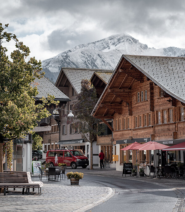 Saanen village centre with typical old chalets, benches, a tree in the foreground and snow-covered mountains in the background.