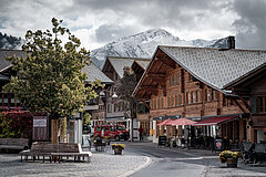 Centre du village de Saanen comprenant de vieux chalets typiques, des bancs et un arbre au premier plan, avec les montagnes enneigées en arrière-plan.