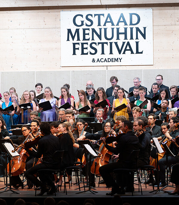 <p>Orchester spielt auf einer Bühne. Im Hintergrund hängt ein Schild mit der Aufschrift Gstaad Menuhin Festival</p>