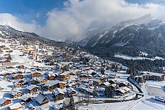 Snow-covered village with clustered chalets, roads and a winter mountain landscape in the background.