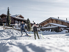 Une skieuse et un skieur de fond sur une bosse lors d’un skills parc de ski de fond. En arrière-plan, des chalets en bois entourés d’arbres sous un ciel bleu.