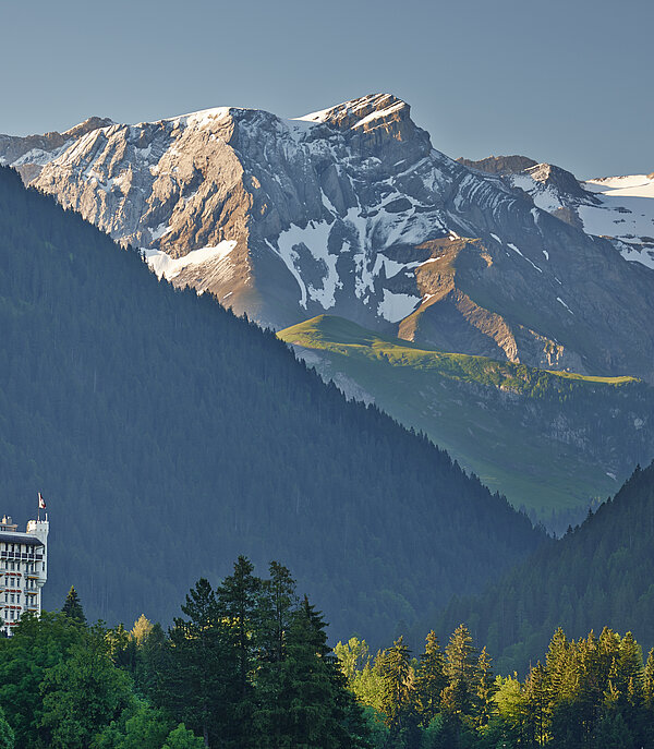 The Hotel Gstaad Palace is built like a castle and overlooks the forest. In the background you can see snowy mountains illuminated by the morning sun.