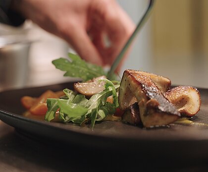Seitliche Nahaufnahme eines schwarzen Tellers, auf dem eine Hand eine Speise anrichtet. Auf dem Teller werden frische Steinpilze mit Rucola und Karotten drappiert.