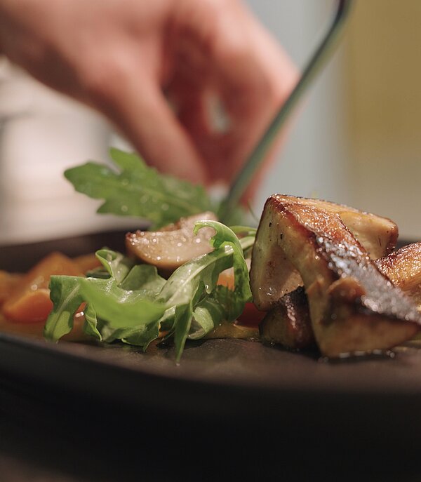 Side close-up of a black plate as a hand arranges a dish with fresh porcini mushrooms, arugula, and carrots.