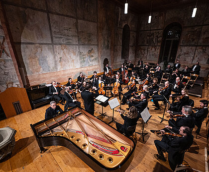 Ein Orchester spielt klassische Musik in einer Kirche mit christlichen Sujets an den Wänden.