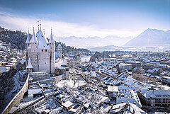 Winterliche Ansicht der Stadt Thun mit verschneiter Altstadt, Schloss Thun auf dem Hügel und Berglandschaft im Hintergrund.