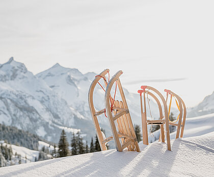  Three wooden sledges stand at the top of the mountain in fresh powdery snow, with white mountain peaks in the background.