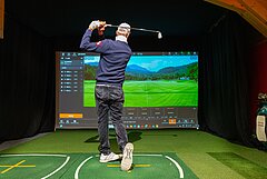 A man plays golf on a green carpet in an indoor facility in front of a screen showing a golf course.