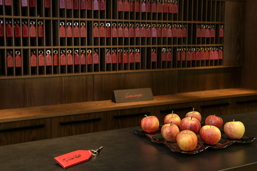 <p>Empfangstheke mit einem Zimmerschlüssel mit rotem Anhänger und einer Schale mit Äpfeln, im Hintergrund Schlüsselbrett mit Zimmerschlüsseln.</p>
