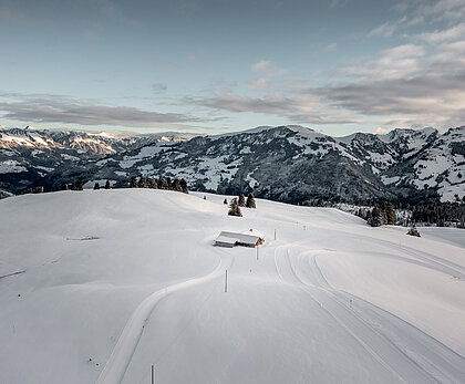 Verschneite Winterlandschaft mit einer Langlaufloipe und einem breiten Winterwanderweg, dahinter eine endlos herrliche Bergkulisse und leicht bewölkter Himmel. 