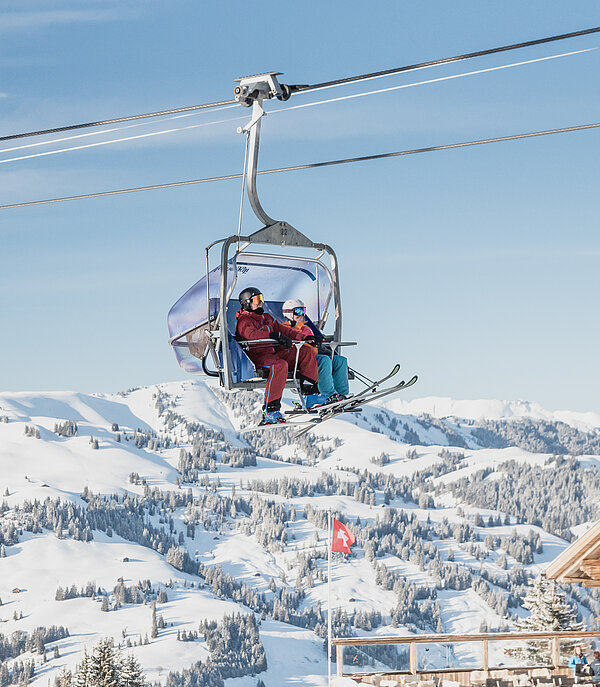 Sessellift mit 2 Skifahrern und verschneiter Aussicht.&nbsp;
