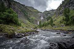  Une imposante chute d’eau se jette dans la vallée. Au premier plan, un ruisseau, derrière, des montagnes.