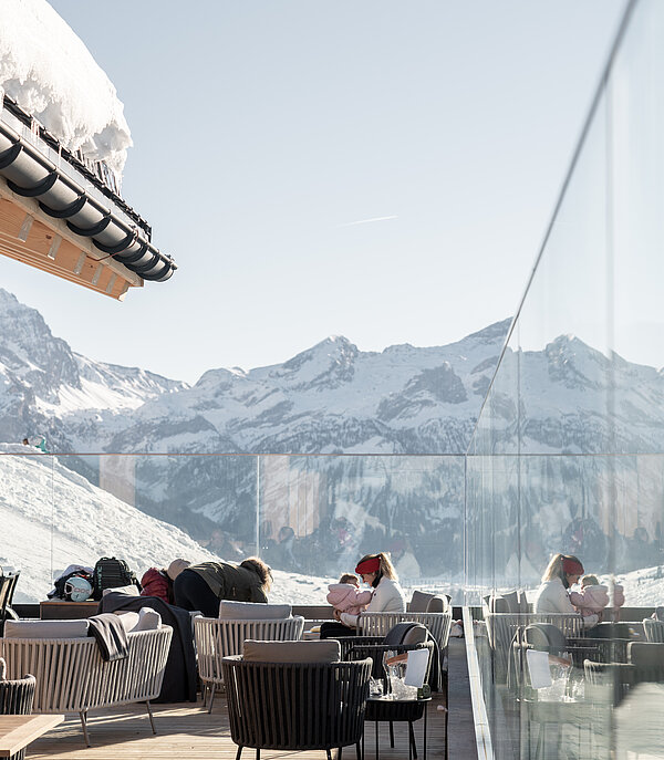 Guests sitting at a mountain restaurant terrace in winter, protected from the wind by glass walls, with a beautiful snowy mountain landscape in the background.
