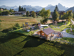 Der Spiellatz mit dem Picknickhaus liegt eingebettet im Grünen und strahlt im Abendlicht vor dem Bergpanorama.