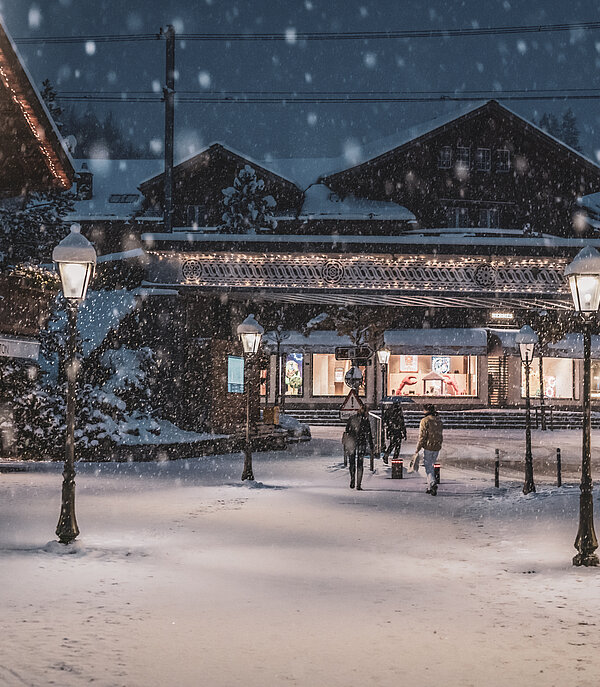 Three people walking along the romantically lit promenade in Gstaad in the evening, with snow falling around them.
