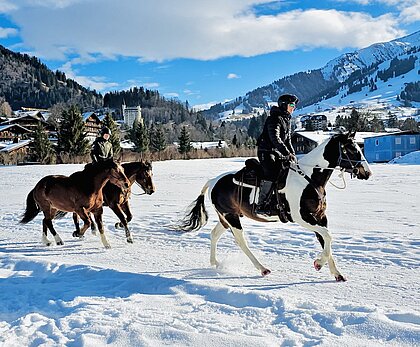 Reiter galoppieren auf Pferden über eine verschneite Wiese vor alpiner Bergkulisse mit dem Dorf Gstaad im Hintergrund.