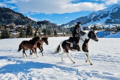 Reiter galoppieren auf Pferden über eine verschneite Wiese vor alpiner Bergkulisse mit dem Dorf Gstaad im Hintergrund.