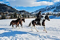 Reiter galoppieren auf Pferden über eine verschneite Wiese vor alpiner Bergkulisse mit dem Dorf Gstaad im Hintergrund.