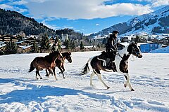 Reiter galoppieren auf Pferden über eine verschneite Wiese vor alpiner Bergkulisse mit dem Dorf Gstaad im Hintergrund.