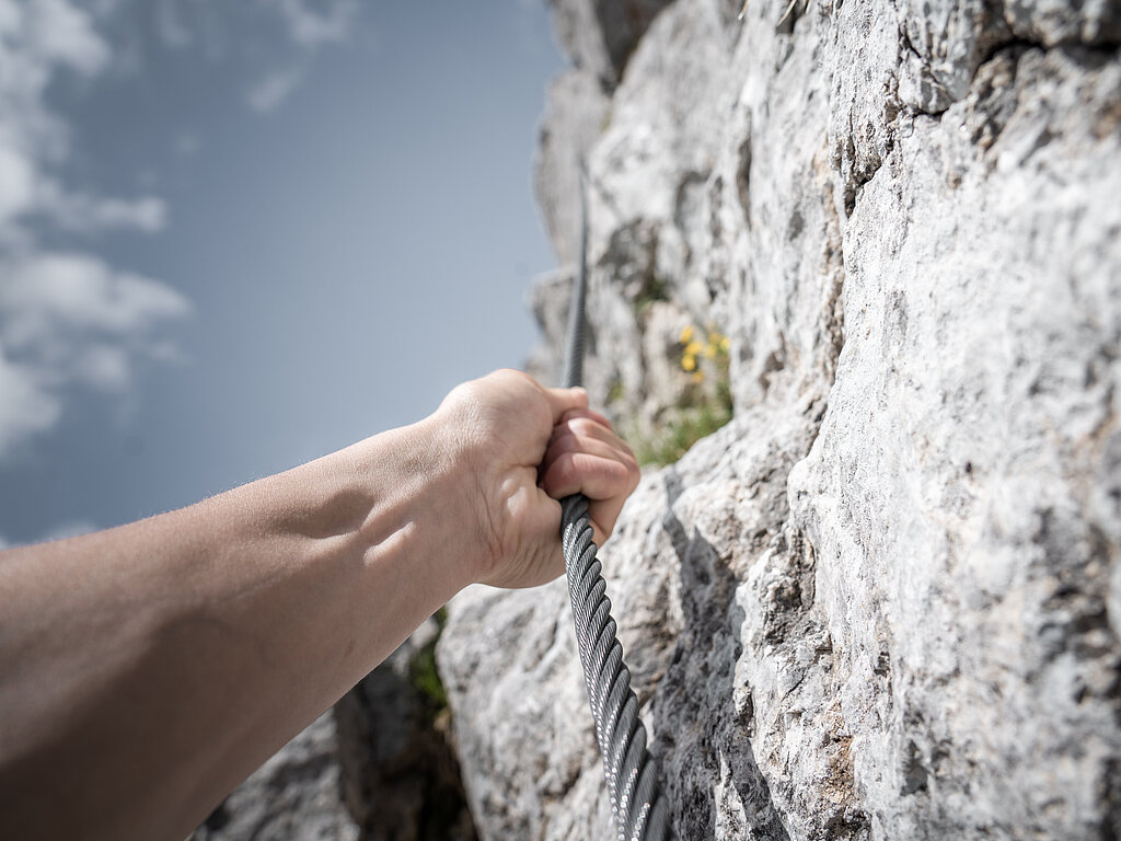 <p>Eine Hand welche sich an einem Stahlseil festhält bei einem Klettersteig.</p>