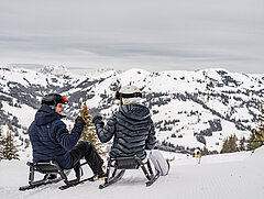 People in dark ski clothes sledding amid a snowy mountain panorama.