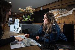 Une employée de l’office du tourisme de Gstaad Saanenland montre quelque chose à une touriste sur une carte panoramique. En arrière-plan, les guichets de l’office de tourisme de Gstaad.